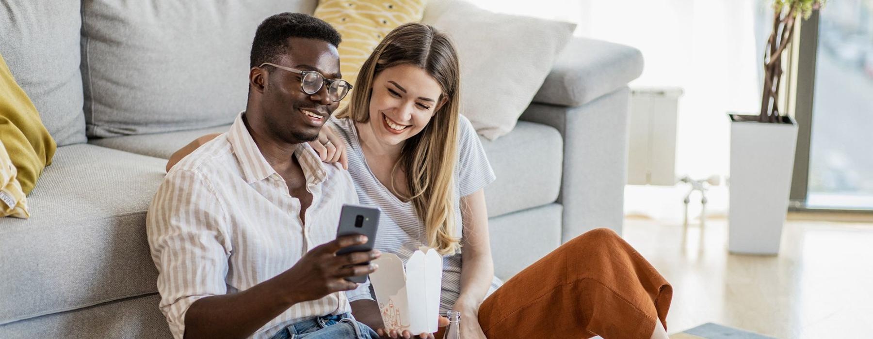 a man and woman with take out, sit against a couch on their living room floor and watch at their cell phone