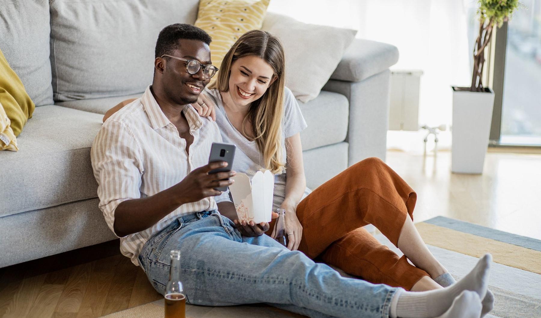 a man and woman with take out, sit against a couch on their living room floor and watch at their cell phone